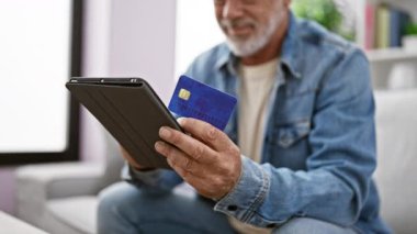 Evde kredi kartı ve tablet tutan sakallı olgun bir adam ev ortamında teknoloji ve finans sunuyor..