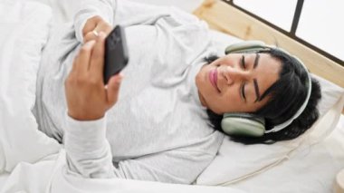 İspanyol kadın, kulaklıkla yatakta dinleniyor, akıllı telefon kullanıyor, modern kapalı mekan eğlencesi tasviri yapıyor..