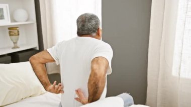 Gri saçlı son sınıf öğrencisi, iyi ışıklandırılmış bir yatak odasında yatağında oturuyor. Sırtını sanki acı çekiyormuş gibi tutuyor..