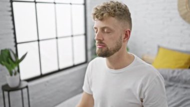Sakallı genç beyaz bir adam modern bir yatak odasında meditasyon yapıyor. Geniş bir penceresi ve arka planda bir bitkisi var..
