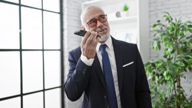 Takım elbiseli olgun işadamı modern ofiste telefonla konuşuyor.