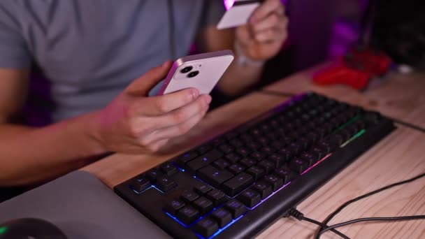 ゲームルームのヒスパニック系男性がスマートフォンとクレジットカードを持ち 夜にオンラインショッピングを準備 — ストック動画