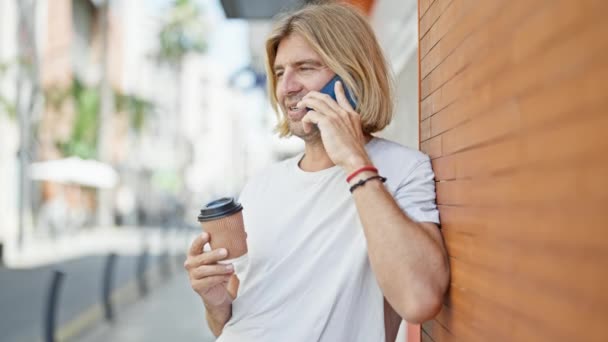 笑顔の金髪の男は 都市の背景に対してコーヒーカップを保持しながら電話を楽しんでいます — ストック動画