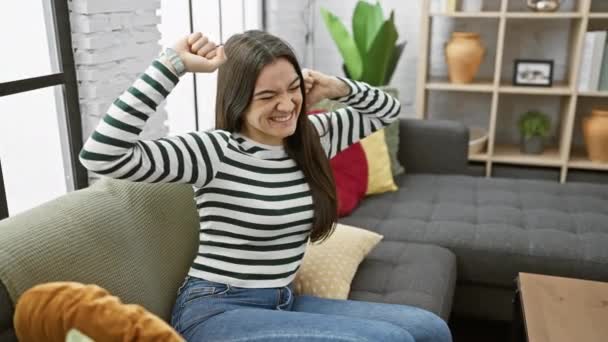 Радостная Молодая Латиноамериканка Сидящая Дома Диване Лопается Взволнованным Жестом Победителя — стоковое видео