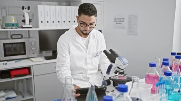 실험실 코트에 집중된 남자는 실험실 환경에서 현미경을 사용하여 샘플을 조사합니다 — 비디오