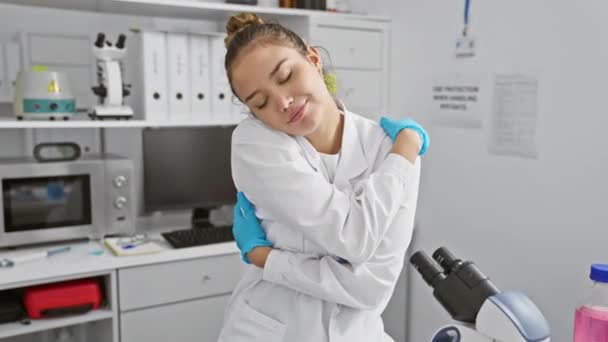 科学者の制服を着た陽気な若いヒスパニック系女性が 研究室で自信を持って抱きしめている 自己愛 そして研究室でのポジティブなバイブを表現する — ストック動画