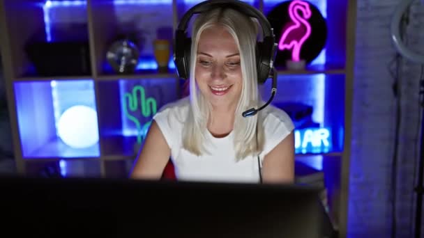 若い金髪のゲーマーの女性が面白い表現で舌を突き出し 完全に陽気なビデオゲームに包まれ 暗いゲームルームで喜びと狂気に幸せを感じる — ストック動画