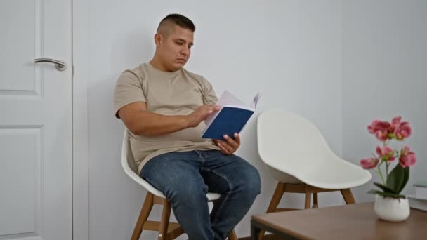 英俊的拉丁男人 沉浸在一本书里 舒服地坐在大堂的椅子上 一个年轻的 放松的 严肃的成年人 他的注意力集中在候车室的内部 — 图库视频影像