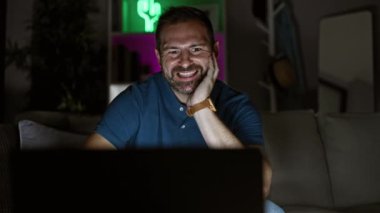 Geceleri rahat, loş bir oturma odasında dizüstü bilgisayar kullanan orta yaşlı bir adam..