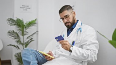 Gözlüklü ve beyaz ceketli sakallı bir adam akıllı telefonunu ve kredi kartını parlak bir muayenehanede kontrol ediyor..