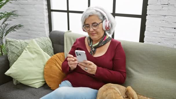 成熟した灰色の髪の女性 屋内ペット犬の隣のソファでスマートフォンを使用してヘッドフォン付き — ストック動画