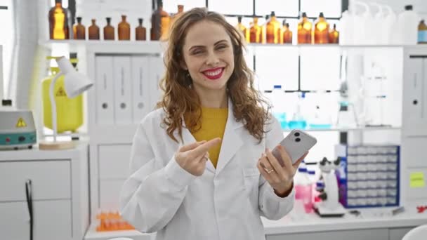 身穿实验室制服的年轻而快乐的女人 拿着智能手机在实验室里欢快地摆弄着姿势和姿势 快乐的表情流露出积极的激情和自信 — 图库视频影像