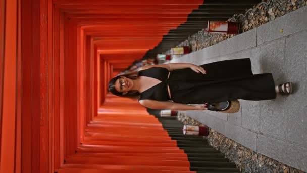 きれいな笑顔と眼鏡で 気楽で楽しい若いヒスパニック女性が 京都のオレンジ色の鳥居の門のカメラに向かって歩いていると自信を持ってポーズします — ストック動画
