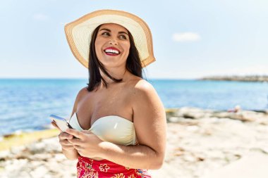 Genç, güzel İspanyol kadın turist deniz kenarında akıllı telefon kullanarak gülümsüyor.