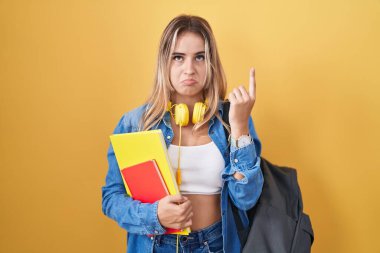 Öğrenci sırt çantası takan, elinde kitapları tutan üzgün ve üzgün görünen, yönünü parmaklarıyla gösteren mutsuz ve depresif genç sarışın bir kadın.. 