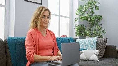 Modern oturma odasındaki koltukta dizüstü bilgisayar kullanan olgun sarışın bir kadın..