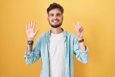 Sarı arka planda dikilen İspanyol dövmeli genç adam yedi numaralı parmaklarıyla gülümseyerek kendinden emin ve mutlu olduğunu gösteriyor.. 