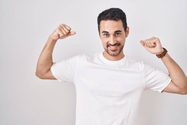 Beyaz arka planda duran yakışıklı İspanyol adam gururla gülümseyen kol kaslarını gösteriyor. fitness konsepti. 