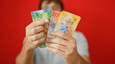 Genç, sarışın, sakallı, yakışıklı adam büyük paralar yatırıyor, İsviçre frangı paralarını sayıyor! çarpıcı bir kırmızı arkaplan karşısında izole