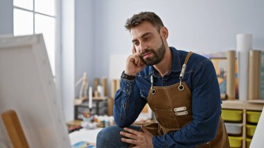 Odaklanmış genç İspanyol erkek sanatçı sanat stüdyosunda oturuyor, yakışıklı sakallı adam ciddi şekilde tuvalden tuvale resim yapıyor.