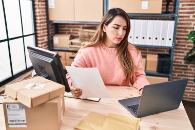 Young beautiful hispanic woman ecommerce business worker using laptop reading paperwork at office