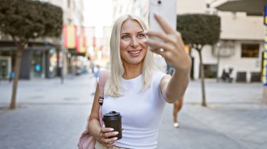 Sarışın genç bir kadın, şehir caddesinde gelişigüzel dikilmiş, akıllı telefonuyla kendinden emin bir şekilde gülümsüyor, telefon elinde, kahvaltıda kahvesini götürürken keyfine bakıyor..