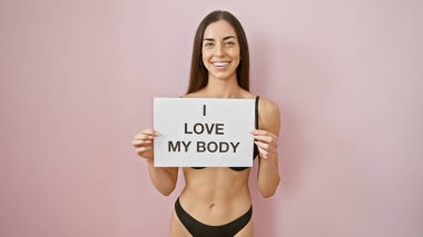 İç çamaşırlı güzel İspanyol bir kadın, kendinden emin bir şekilde şehvetli sabahının tadını çıkarıyor, gururla 'Vücudumu seviyorum' afişini pembe arka planda tutuyor, neşeli ve pozitif bir mesaj yayıyor.