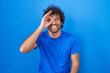 Mavi arka planda dikilen İspanyol genç adam el hareketleriyle gülümsüyor ve mutlu yüzüyle parmaklarının arasından bakıyor.. 