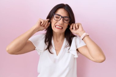 Orta yaşlı İspanyol kadın gündelik beyaz tişört giyiyor ve yüksek sesli müzik sesi için kulakları parmaklarıyla kaplayan gözlük takıyor. sağır kavramı. 