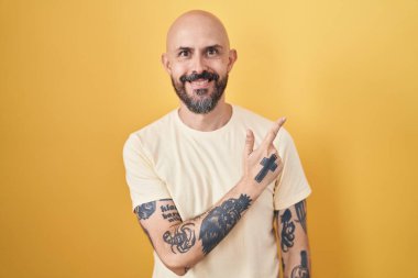 Sarı arka planda dikilen İspanyol dövmeli adam. Yüzünde gülücükler, mutlu ve doğal bir ifadeyle el ve parmak uçlarını gösteren bir gülümseme. 