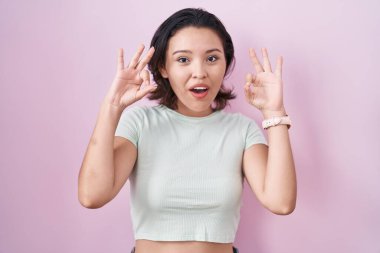 Pembe arka planda duran Latin kökenli genç kadın şaşırmış ve şok olmuş görünüyor. Tamam sembolünü parmaklarıyla yapıyor. çılgın ifade 