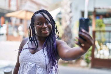 Afro-Amerikalı kadın gülümsüyor. Sokakta akıllı telefonun yanında özçekim yapıyor.