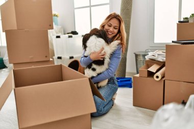 Beyaz bir kadın yeni evinde yerde oturmuş köpeğe sarılıyor.