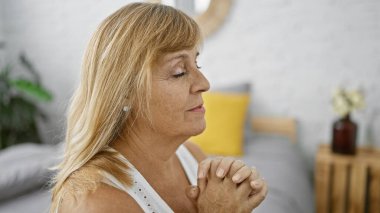 Orta yaşlı bir kadın, güzel bir sarışın, inancı ve umuduyla dua eden rahat bir şekilde yatağında oturuyor, rahatlıyor ve rahat bir yatak odasında dinleniyor.