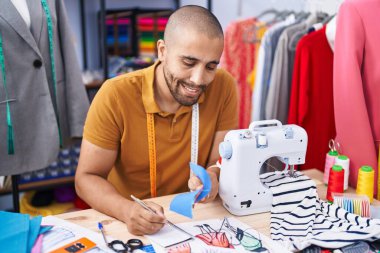 Young latin man tailor writing on notebook holding cloth at atelier