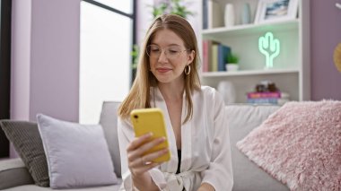 Kanepede oturmuş akıllı telefon kullanan genç sarışın kadın evde gülümsüyor.