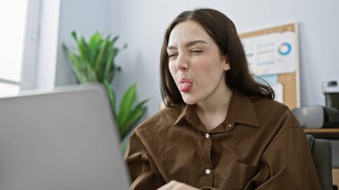 Young caucasian woman making a playful face in a modern office setting, depicting casual workday life. clipart