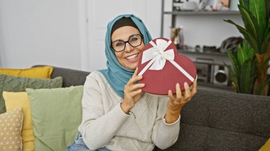 Başörtüsü takan gülümseyen olgun bir kadın rahat bir oturma odasında kalp şeklinde bir hediye kutusu tutuyor..
