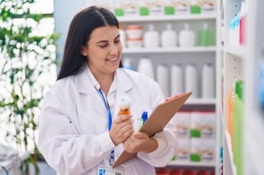 Genç ve güzel İspanyol kadın eczacı elinde ilaç şişesiyle eczanede doküman okuyor.