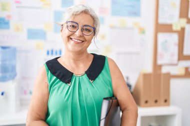 Orta yaşlı, gri saçlı, iş kadını. Ofiste elinde bir not panosuyla gülümsüyor.