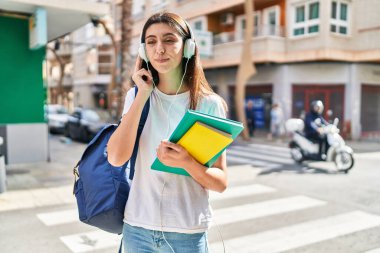 Young beautiful hispanic woman student holding books using smartphone at street