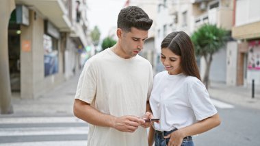 Gündelik giysiler içinde bir kadın ve bir adam sakin bir caddede bir anı paylaşıyorlar, akıllı telefona birlikte bakıyorlar..