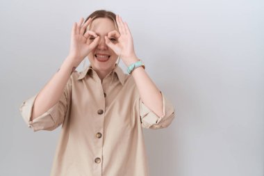 Kafkasyalı genç bir kadın günlük gömlek giyiyor dürbün gibi jest yapıyor, dili dışarı çıkarıyor, gözleri parmaklarından bakıyor. çılgın ifade. 