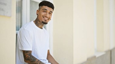 Neşeli genç latin adam kendine güveni ve mutluluğu yayıyor. Şehirli bir sokakta rahatça otururken geniş bir gülümseme.