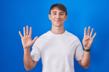 Beyaz, sarışın bir adam mavi arka planda dikiliyor ve dokuz numaralı parmaklarıyla işaret ediyor kendinden emin ve mutlu bir şekilde gülümsüyor.. 