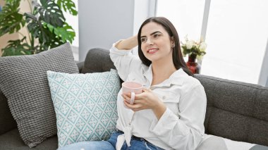 Rahat İspanyol kadın rahat bir oturma odasında rahat bir kanepede kahvesini içiyor..