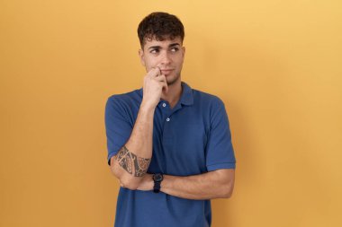 Sarı arka planda, elleri çenesinde soru sormayı düşünen genç İspanyol bir adam. Düşünceli bir ifade. Düşünceli bir yüzle gülümsüyor. şüphe kavramı. 