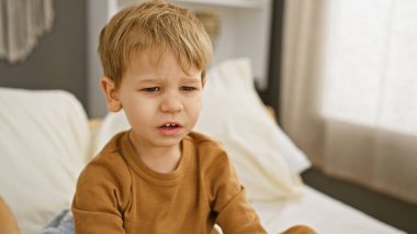 Sarışın bir çocuk evde bir yatakta otururken üzgün görünür..