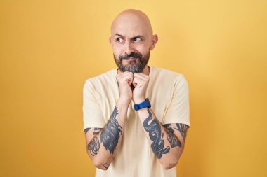 Sarı arka planda dikilen İspanyol dövmeli adam elleri çenesinde gergin ve heyecanlı bir şekilde yan tarafa bakıyor. 