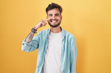 Sarı arka planda dikilen İspanyol dövmeli genç bir adam. El ve burunla işaret ediyor. Neşeli gülümsüyor. güzellik konsepti 